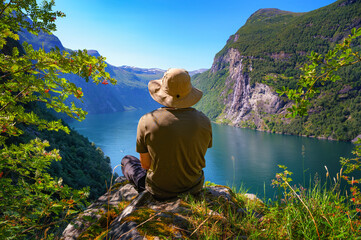 Hiker wearing a straw hat sits on a hilltop with views of Geiranger Fjord and Seven Sisters Waterfall in Norway.
