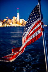 Night view of Manhattan skyline with flag. American flag waves gently as boats glide through water, showcasing Manhattan's illuminated skyline during the night.