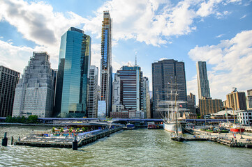 Fototapeta premium Manhattan skyline with waterfront park. Skyline of Manhattan featuring modern skyscrapers and a sailboat near the bustling waterfront park during daylight.