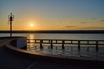 A minimalist sunset scene with a bright orange sky, calm sea, and a red navigation light standing against the horizon, casting long reflections on the water in Gdynia, Poland.