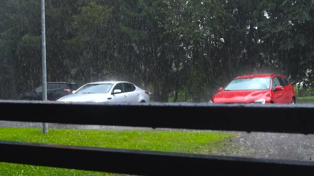 Slow motion video of parked gary and red cars under heavy rainfall during monsoon season at summer or autumn cloudy day with green grass and trees visible in a residential neighbourhood. Wooden rails.