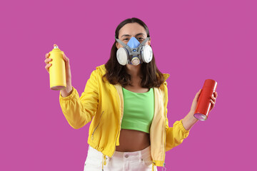 Young woman in respirator with spray paint cans on purple background