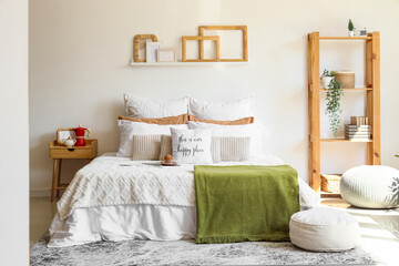 Big comfortable bed with muffin, bedside table, shelving unit and poufs in interior of bedroom