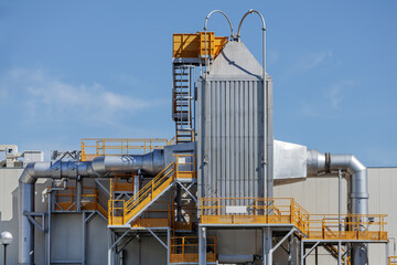 Housing of towering rectangular filter system dominates industrial production facility scene, featuring piping network connected to central tower surrounded by bright orange stairways and platforms.