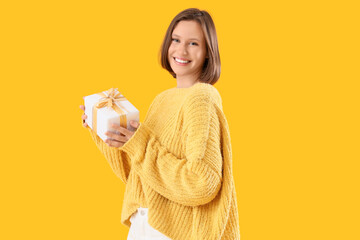 Young woman in warm sweater with Christmas gift box on yellow background