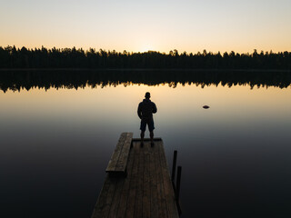 Silhouette of a person standing on a wooden pier, enjoying the tranquil sunrise over a still...