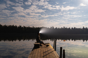 Man sitting on a wooden pier by a tranquil lake, shining a powerful headlamp beam towards the...