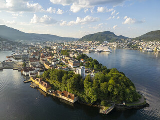Aerial view of a peninsula with lush greenery and historic buildings jutting into the still, dark waters, framed by distant mountains, Bergen, Vestland, Norway.
