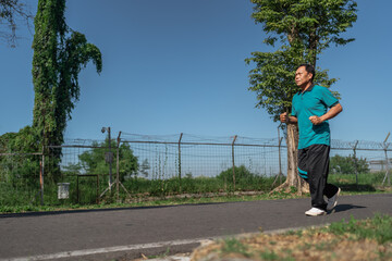A man is jogging steadily on a lovely pathway, surrounded by lush greenery and clear blue skies