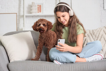 Teenage girl in headphones with Poodle dog using mobile phone on sofa at home