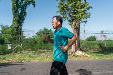 An elderly man jogs energetically in a peaceful park surrounded by beautiful trees on a sunny day