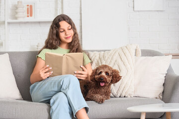Teenage girl with Poodle dog reading book on sofa at home