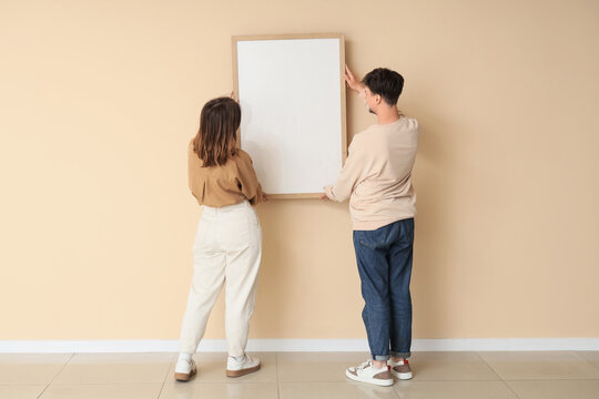 Young couple hanging blank frame on beige wall, back view