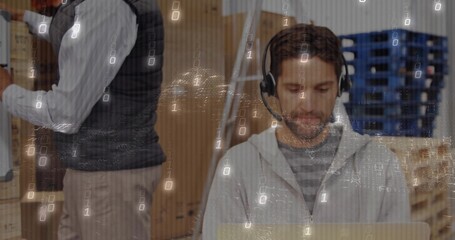 Typing IT specialist on silver laptop with headset in warehouse area, coworker stacking boxes