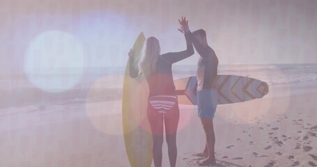High-fiving adult surfers wearing wetsuits on beach sand at sunrise, with surfboards, footprints