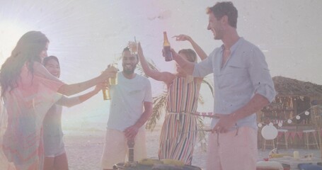Friends raising beer bottles and toasting barbecue on sunset beach, with grill and string lights