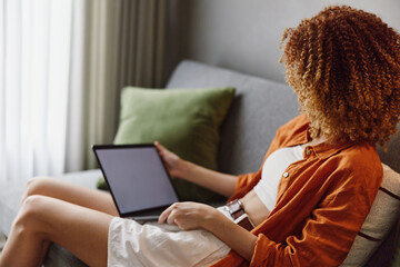 Young woman with curly hair in orange shirt using laptop, relaxed at home, showcasing modern lifestyle and technology in a cozy indoor setting