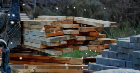 Forklift forks supporting pile of planks near stacked blocks in supply yard, snowflakes falling