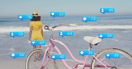 Standing woman wearing yellow dress and sunhat at beach, with pink bicycle, floating notifications