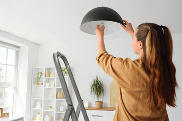 Young woman screwing light bulb into hanging black lamp at home, back view