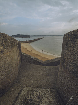 View of the golden beach is framed by ancient stone walls under a vast, cloudy sky, with the sea stretching to the horizon, Saint-Malo, Brittany, France.