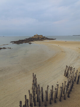View of weathered wooden posts stand firmly on the expansive sandy beach leading to a distant stone fort under a muted sky, Saint-Malo, Brittany, France.