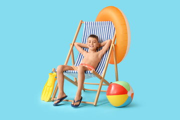 Cute little boy with beach accessories sitting on deckchair against blue background