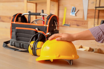 Male hand with hardhat, hearing protectors and tool bag on table in workshop