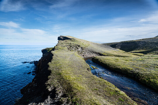 View of rugged cliffs and a narrow stream meet the vast, deep blue ocean under a clear sky, creating a serene yet dramatic landscape, Ketubj&Atilde;&para;rg, Iceland.