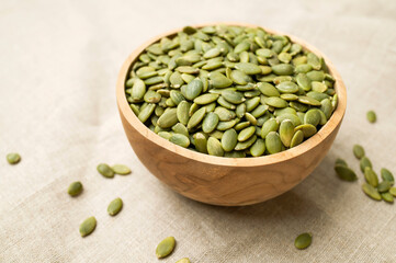 Raw peeled green pumpkin seeds in wooden bowl on natural linen fabric background top view. Food...