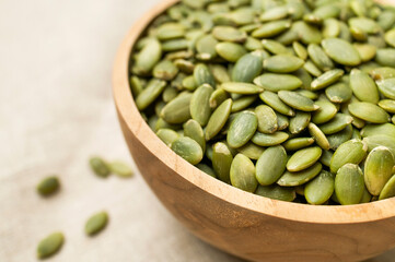 Raw peeled green pumpkin seeds in wooden bowl on natural linen fabric background top view. Food...