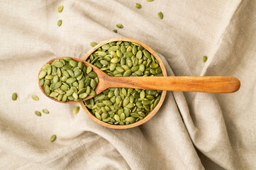 Raw peeled green pumpkin seeds in wooden bowl and a spoon on natural linen fabric background top...