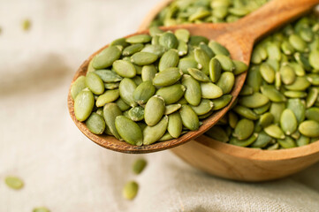 Raw peeled green pumpkin seeds in wooden bowl and a spoon on natural linen fabric background top...