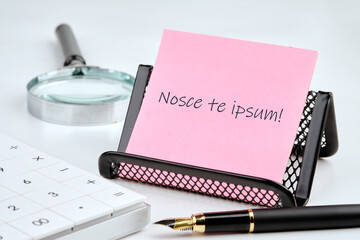Latin proverb NOSCE TE IPSUM (know yourself) on a pink sticker with office supplies on a white...