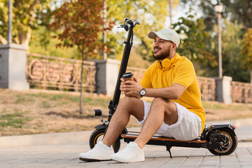 Handsome young man with cup of coffee sitting on electric scooter at city street