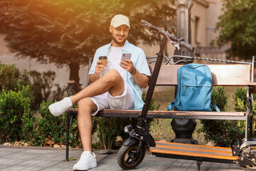Handsome young man with coffee, mobile phone and electric scooter sitting on bench at city street