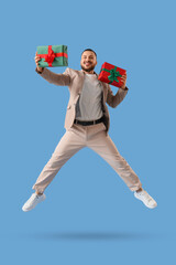 Handsome young man with Christmas gifts jumping on blue background