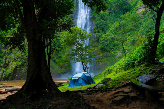 View of a tent pitched on the verdant ground near a towering waterfall, with the scene framed by lush trees in Hohoe, Volta Region, Ghana.