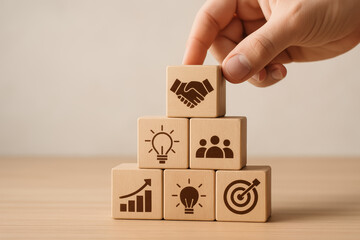 Hand placing a block on a pyramid of wooden cubes featuring business symbols like growth, collaboration, and innovation.