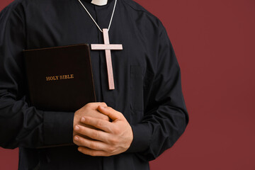 Young priest with wooden cross and Bible on red background, closeup