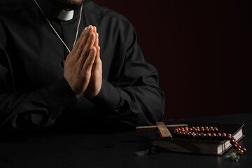 Young priest with wooden cross, beads and Bible praying at table on red background, closeup