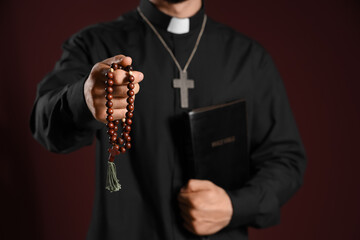 Young priest with beads, Bible and wooden cross on red background, closeup