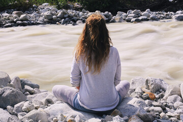Caucasian young adult woman sitting cross legged on rocky riverbank facing flowing water, long wavy...