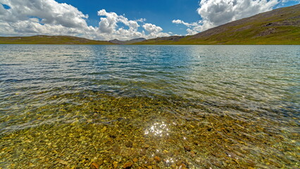 View of glimmering sunlight dancing on the clear, shallow lake waters revealing the rocky bed with distant mountains under a partly cloudy sky, Deosai National Park, Gilgit Baltistan, Pakistan.