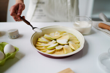 Unknown woman sprinkling sugar over sliced apples in baking dish, preparing dessert in kitchen,...
