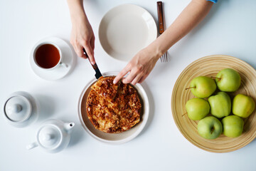 Top down of woman cutting pie with knife at table, hands visible near plate with green apples, cup of tea and teapots arranged on surface, top view composition