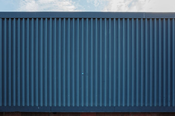 a modern industrial building exterior, featuring a blue corrugated metal wall with a repeating vertical line pattern. A red brick foundation is visible at the bottom and a blue sky at the top.