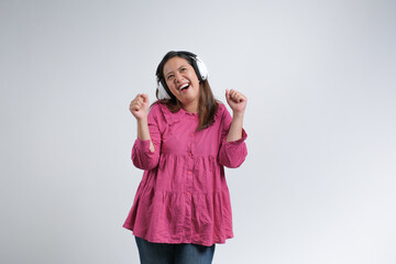 A cheerful young woman wearing a pink blouse and jeans enjoying music with wireless headphones,...