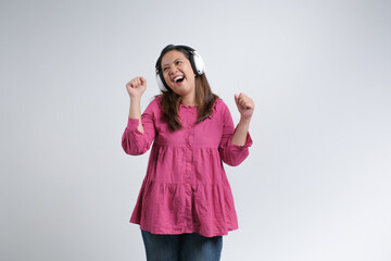 A cheerful young woman wearing a pink blouse and jeans enjoying music with wireless headphones,...