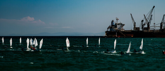 A scene from a sailboat race at sea. Numerous small Optimist-class sailboats are visible, each with...
